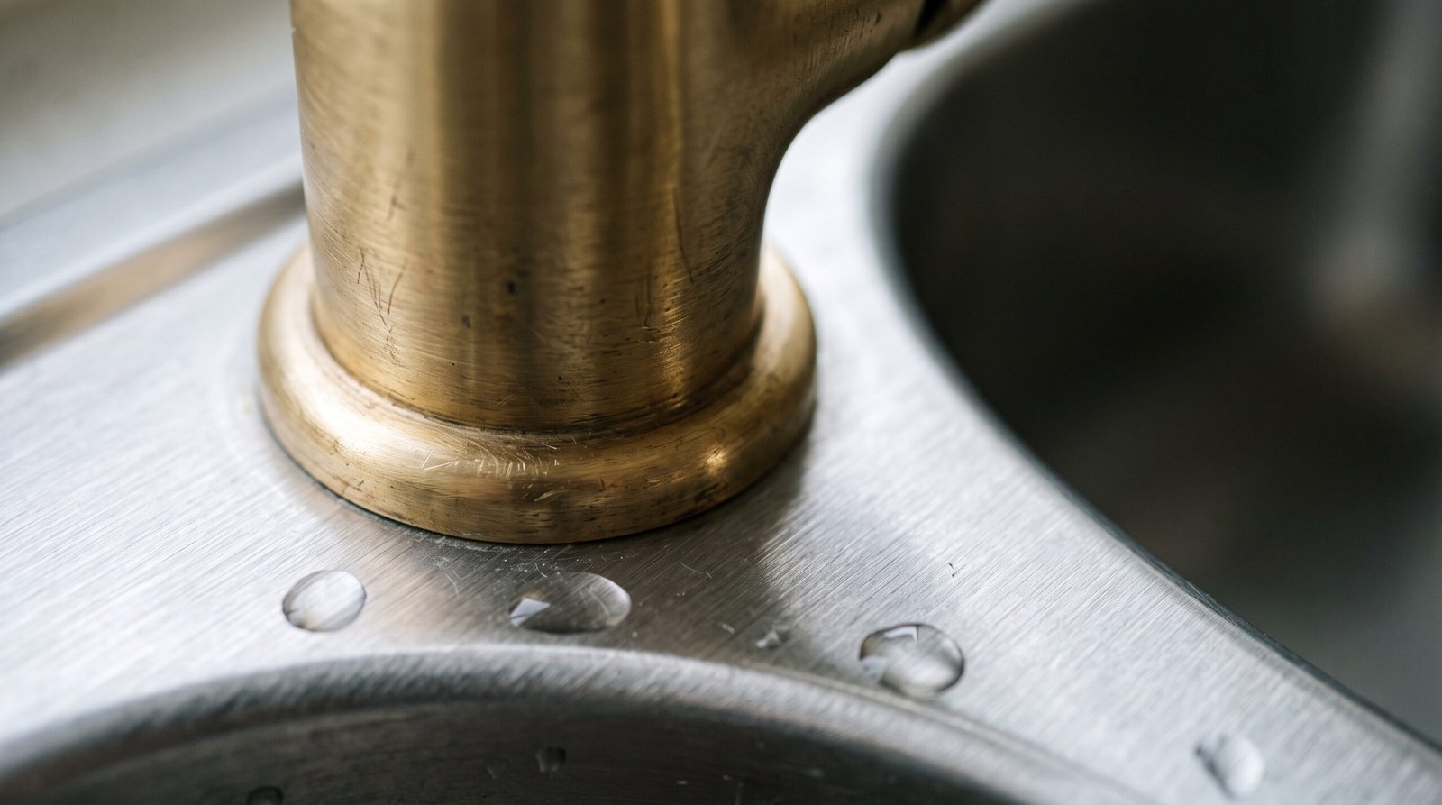 Close view of the base of an unlacquered brass kitchen faucet where it meets the thin brushed stainless steel deck of a sink