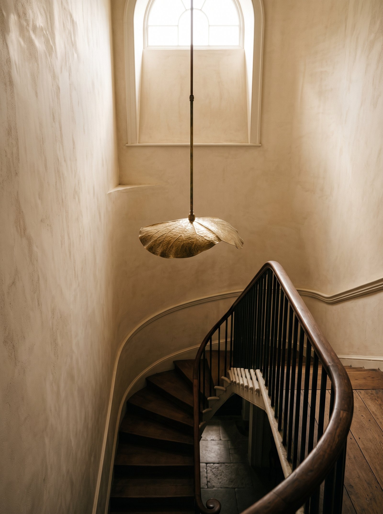 A brass leaf pendant hanging on a long stem in a plastered stairwell, lit from an arched window above the turn of the stairs