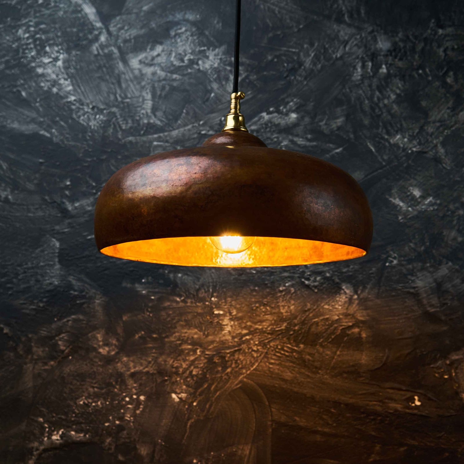 Oxidized copper dome pendant light glowing warm against a dark plaster wall